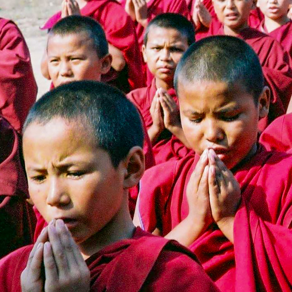 Children in maroon robes with hands pressed together in a prayer gesture.
