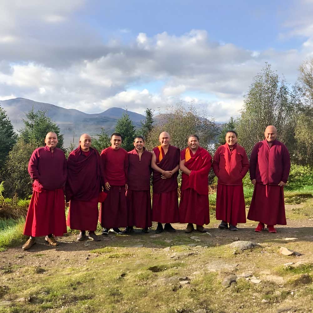A line of monks in maroon robes standing outdoors on a grassy hill with mountains in the background.